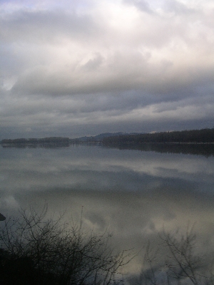 [The sky is nearly 100 percent covered in clouds, but there is color variation in them and this is reflected in the water. The image is nearly all sky and water with only a thin line of the far shore visible.]